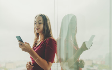 Wall Mural - Smiling attractive caucasian businesswoman using smartphone near glass wall with reflection, beautiful adult woman entrepreneur in red dress standing near window of office skyscraper with cellphone