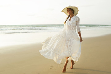 Woman wearing beautiful white dress is walking on the beach during sunset