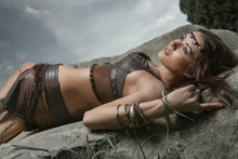 Young Beautiful Woman Dressed In A Leather Wear Looking Like An Amazon Or Warrior Lying On A Big Stone. Dramatic Beauty Shot