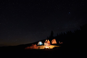 Wall Mural - Night camping in the mountains. Couple tourists have a rest at a campfire near two tents under starry sky at night. Long exposure