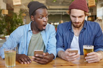 Portrait of two interracial friends sitting at cafeteria drinking beer playing games online using modern gadgets. Surprised black hipster looking at mobile phone of bearded male seeing something