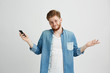© Cookie Studio - Portrait of young handsome guy holding smart phone shrugging looking at camera over white background.