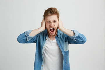 Wall Mural - Angry rage young man with beard shouting screaming closing ears over white background.