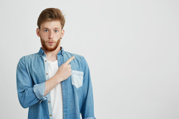 Wall Mural - Portrait of surprised young handsome man looking at camera pointing finger up over white background.