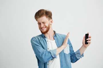 Wall Mural - Displeased young handsome man showing stop sign to phone in another hand looking at camera resisting over white background.