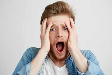 Wall Mural - Frightened sad upset young man looking at camera with opened mouth holding head with hands over white background.