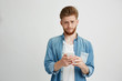© Cookie Studio - Dissapointed upset young man with beard holding smart phone looking at camera over white background.