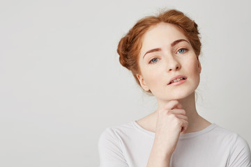 Wall Mural - Portrait of young pretty girl with red hair looking at camera touching chin over white background.