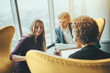 © skyNext - Attractive smiling businesswoman is showing screen of her gadget to her boss and female colleague during work meeting while sitting on yellow armchair in office interior next to window of skyscraper