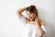 © shurkin_son - Beauty and makeup concept. Beautiful tender young European woman model wearing white top with open shoulders and black choker, looking down and adjusting her hairstyle while posing in studio