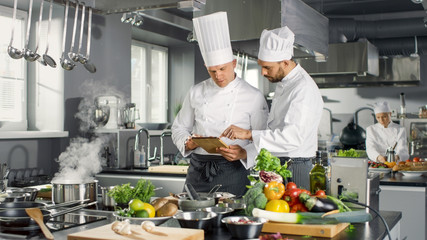 Two Famous Chefs Discuss Their Video Blog while Using Tablet Computer. They Work on a Big Restaurant Stainless Steel Professional Kitchen.