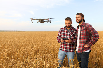 Compact drone hovers in front of two hipster men. Quadcopter flies near farmer and agronomist exploring harvest with innovative technology taking aerial photos and videos from above it wheat field