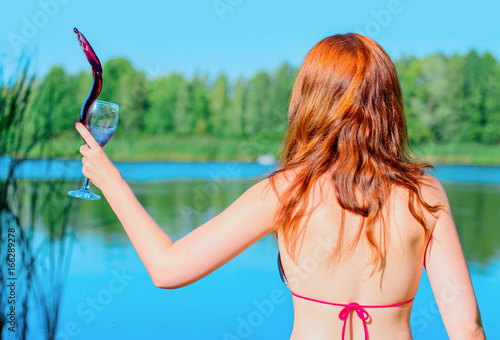 Fotografie, Obraz Red hair young woman in swimsuit rest near blue water with glass of red wine