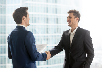 Happy satisfied business partners shaking hands after concluding contract for services on city building background, two smiling businessmen seal good deal, binding bargain with firm strong handshake