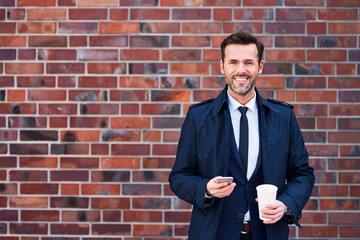 Joyful middle-aged business executive holding phone and coffe in front of a brick wall