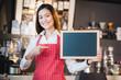 Asian female barista wear red apron point at blank blackboard coffee menu at counter bar with smile face,cafe service concept,Leave space for adding your text