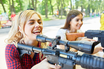 Portrait of two beautiful women aiming with big rifle at outdoor game tent in amusement park