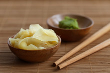Pickled Ginger Slices In Wooden Bowl On Bamboo Mat Next To Chopsticks And Wasabi Paste In A Bowl.