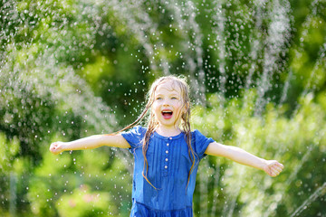 Wall Mural - Adorable little girl playing with a sprinkler in a backyard on sunny summer day. Cute child having fun with water outdoors.