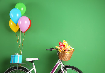 Bicycle with basket of flowers, gift and balloons on color background