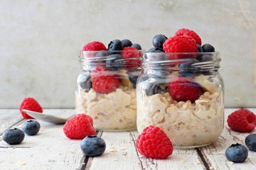 Wall Mural - overnight oats with fresh blueberries and raspberries in jars on a rustic white wood background