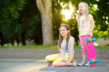 Wall Mural - Two pretty little girls learning to skateboard on beautiful summer day in a park. Children enjoying skateboarding ride outdoors.