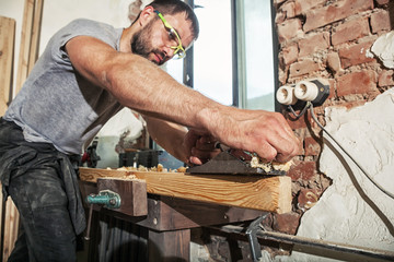 Wall Mural - A dark-haired man with a beard and protective green glasses treating a wooden bar with a black jack plane, work with wooden