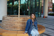 © shurkin_son - Urban portrait of fashionable teenage girl wearing full skirt and denim jacket sitting alone on round-shaped wooden bench waiting for bus to get to city center and meet her friends for a walk