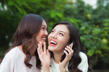 Wall Mural - Young woman whispering into cheerful friend's ear while on call