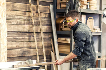 Wall Mural - A young man with dark hair, a beard, wearing goggles and a construction uniform holds a wooden long beam and cuts it on a circular saw in the workshop