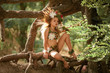 Beautiful little girl in image of nymph dryad sits on tree roots in magical forest .