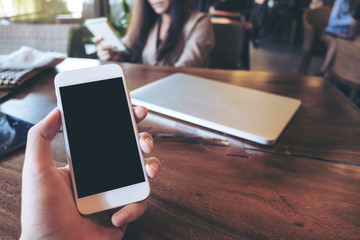 Canvas Print - Mockup image of a man's hand holding white mobile phone with blank black screen in vintage cafe with laptop on wooden table and blur asian business woman in background