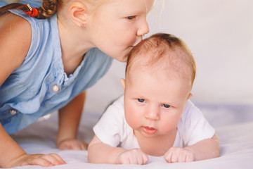 Wall Mural - Lifestyle closeup portrait of cute white Caucasian girl sister kissing little baby, lying on bed indoors. Older sibling with younger brother newborn. Family love bonding together concept.