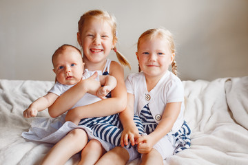 Wall Mural - Lifestyle portrait of cute white Caucasian girls sisters holding little baby, sitting on bed indoors. Older siblings with younger brother sister newborn. Family love bonding together concept.