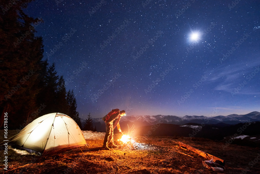 Male tourist have a rest in his camp at night near campfire and tent ...