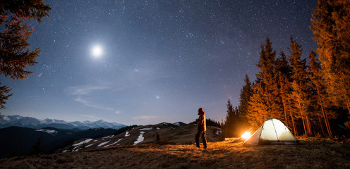 Male tourist have a rest in his camp near the forest at night. Man standing near campfire and tent under beautiful night sky full of stars and the moon, and enjoying night scene. Panoramic landscape