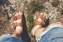 Top View Of Male Legs In Brown Leather Sandals And Blue Jean Shorts, Standing On A Rocky Trail