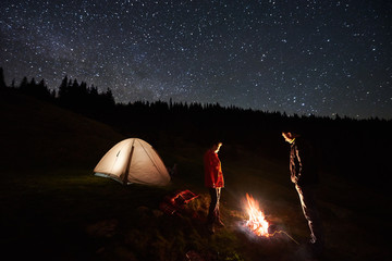 Wall Mural - Night camping. Couple tourists standing at a campfire near illuminated tent under starry sky at night. Long exposure