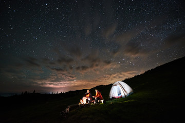 Wall Mural - Night camping. Romantic couple tourists enjoying at a campfire near illuminated tent under beautiful starry sky at night. Long exposure