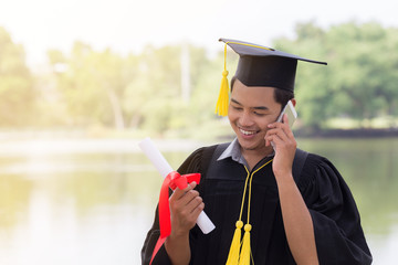 Happy graduated young man in cap and gown talking with parent in mobile phone and looking certificated in hand with nature background