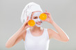 Young woman with facial mask holding slices of orange on gray background.