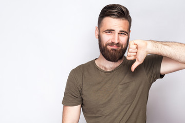 Wall Mural - Portrait of unsatisfied bearded man with thumbs down and dark green t shirt against light gray background. studio shot.  .