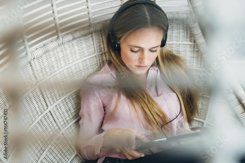 Fotografija Woman in wicker chair listening to music with headphones, reading on a tablet co