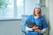 © Zdenka - Young female medical doctor in blue uniform standing in a hospital consulting room, smiling looking into camera. Woman is holding folder and pen, stethoscope is around her neck.