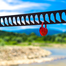A Single Heart Shaped Red Padlock Symbolizing Love Hanging On The Bridge Handrails With Blurred River, Greenery, Mountains And Blue Sky In Background