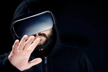 Poster - Young hipster bearded man in black hooded top, wearing virtual reality goggles. Black background studio VR concept