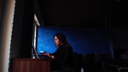 Wall Mural - Educated and elegant businesswoman loves work and makes plans or writes reports on laptop keyboard, smiles and looks carefully at gadget's screen, sits at table in modern cafe on winter day. Beautiful