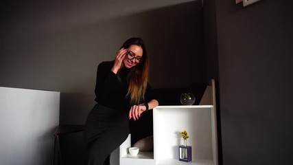 Wall Mural - Pretty woman in beautiful glasses sets up smart watch and uses it for entertainment, smiles and looks at small screen of fashionable gadget, crouching on cube in stylish cafe on winter day. Beautiful