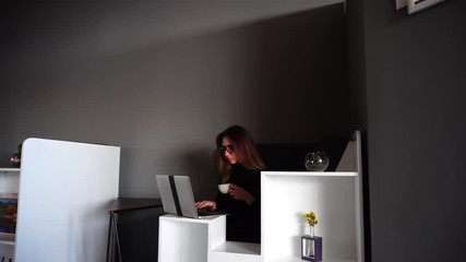 Wall Mural - Charming girl reviewer in Internet newspaper writes for laptop article and dials on keyboard, slowly drinks cup of tea or coffee and sits at desk in office of modern office. Beautiful European-looking