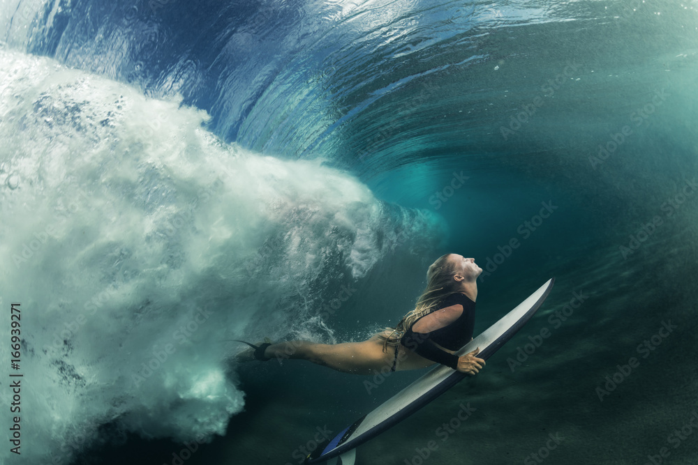 A blonde surfer girl underwater doing duck dive holding surfing board left behind air bubbles in blue water background under big ocean wave - obrazy, fototapety, plakaty 